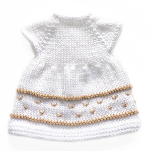 May include: A white knitted dress with short sleeves. The dress features two rows of tan-colored decorative stitching with small heart-shaped accents. The dress is made of a soft, textured yarn.