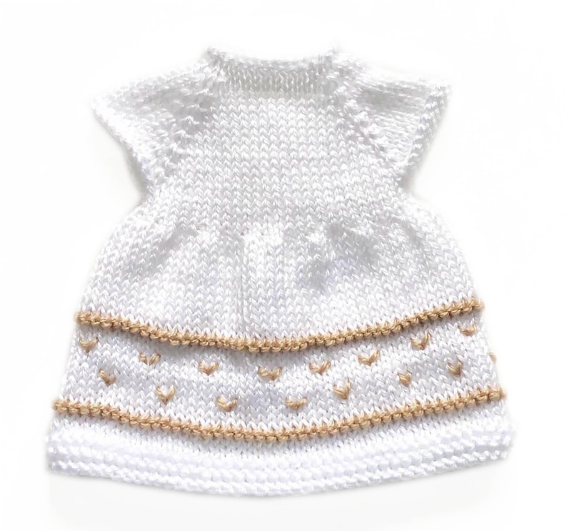 May include: A white knitted dress with short sleeves. The dress features two rows of tan-colored decorative stitching with small heart-shaped accents. The dress is made of a soft, textured yarn.