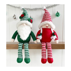 May include: Two knitted Christmas gnomes, one in green and white stripes and one in red and white stripes, sitting on a shelf. Both gnomes have white beards and white pom-poms on their hats.