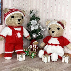 May include: Two knitted teddy bears, one in red dungarees and a striped hat, the other in a red and white dress and hat. A small Christmas tree and wrapped gifts are in the background. The bears are part of a Christmas theme.