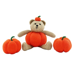 May include: A knitted teddy bear wearing an orange pumpkin costume, with two additional orange pumpkins. The bear has tan arms and head, with a small pumpkin hat. The pumpkins have green stems. The background is white.