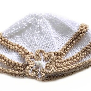 May include: A white knitted baby hat with a tan trim and a flower accent. The hat is made of soft yarn and features a delicate pearl in the center of the flower. The hat is designed for infants and toddlers.
