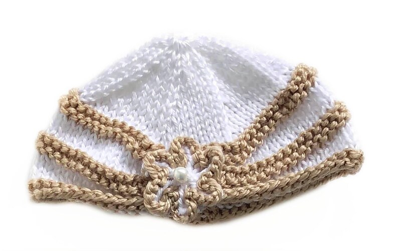 May include: A white knitted baby hat with a tan trim and a flower accent. The hat is made of soft yarn and features a delicate pearl in the center of the flower. The hat is designed for infants and toddlers.