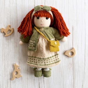 May include: A knitted doll with red hair, dressed in a green cardigan, dress, and shoes. The doll has a yellow handbag and a headband. Wooden toys, including a rocking horse and bird, are placed around the doll.