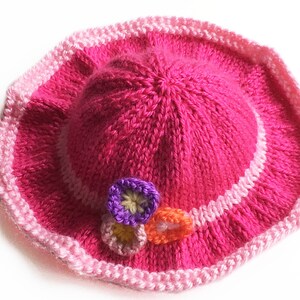 May include: A bright pink knitted hat with a wide brim. The brim is trimmed with a lighter pink and white stripe. Three small, colorful flower appliqu&eacute;s are attached to the side of the hat. The hat is a handmade item.