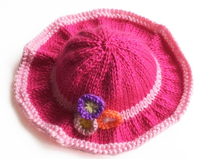 May include: A bright pink knitted hat with a wide brim. The brim is trimmed with a lighter pink and white stripe. Three small, colorful flower appliqu&eacute;s are attached to the side of the hat. The hat is a handmade item.