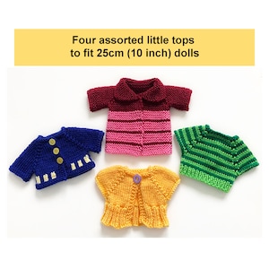 Pode incluir: Quatro tops de malha em diferentes cores e padrões, projetados para bonecas de 25 cm (10 polegadas). Os tops incluem uma camisa listrada rosa e marrom, um cardigan azul com botões amarelos, um cardigan amarelo com um botão roxo e uma camisa listrada verde e branca.