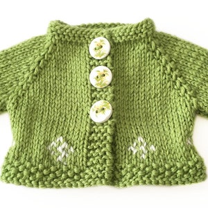 May include: A knitted green cardigan with three floral-patterned buttons and white embroidered accents. The short-sleeved sweater is made of a soft, textured yarn. The garment is designed for a baby or toddler.