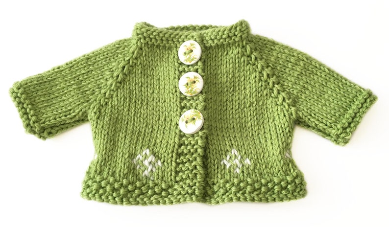 May include: A knitted green cardigan with three floral-patterned buttons and white embroidered accents. The short-sleeved sweater is made of a soft, textured yarn. The garment is designed for a baby or toddler.