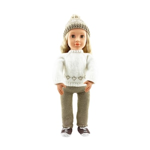 Puede incluir: Una muñeca de pelo rubio largo que lleva un gorro de punto beige, un suéter blanco de manga larga con un sutil patrón de diamantes en la cintura, pantalones de punto marrones y zapatos de punto marrones y blancos. Tiene piel clara.
