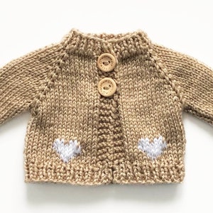 May include: A tan knitted cardigan with two wooden buttons and two white heart appliqu&eacute;s. The miniature sweater has long sleeves and a ribbed collar and cuffs. The garment is designed for a baby or small doll.
