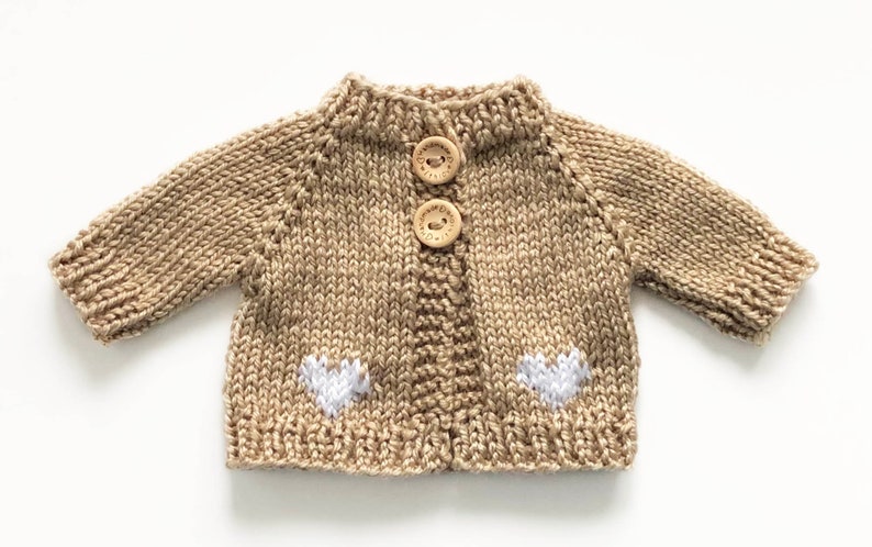 May include: A tan knitted cardigan with two wooden buttons and two white heart appliqu&eacute;s. The miniature sweater has long sleeves and a ribbed collar and cuffs. The garment is designed for a baby or small doll.