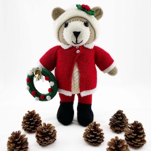 May include: A knitted teddy bear wearing a red Santa Claus outfit with a white-trimmed jacket and hat. The bear holds a green wreath with red and white accents and a small bell. Brown pine cones are scattered around the bear.