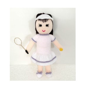 Könnte beinhalten: Handgestrickte Puppe, die eine Tennisspielerin darstellt. Die Puppe trägt ein weißes Tennis-Kleid mit lila Besatz, einen weißen Visor und weiße Schuhe mit lila Schleifen. Sie hält einen Tennisschläger und hat schwarzes Haar.