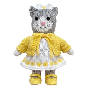 May include: A knitted cat doll featuring gray fur, black eyes, and a pink nose. The doll wears a white dress with a yellow heart pattern along the hem, a yellow cardigan with a tie front, a white bow on its head, and yellow shoes with straps.
