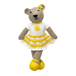 Puede incluir: Un oso de peluche tejido con un vestido blanco con una falda amarilla y un adorno floral amarillo. El oso tiene un cuerpo marrón, ojos negros y zapatos amarillos. El oso mide aproximadamente 30 cm de alto.