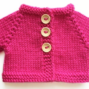 May include: A vibrant fuchsia-colored knitted cardigan with three wooden buttons. The short-sleeved sweater is hand-knitted with a visible stitch pattern. The collar and cuffs are neatly finished. A charming, handmade garment.