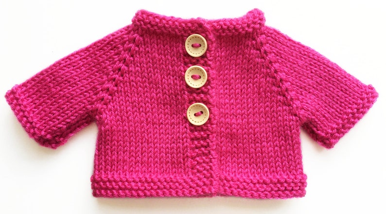 May include: A vibrant fuchsia-colored knitted cardigan with three wooden buttons. The short-sleeved sweater is hand-knitted with a visible stitch pattern. The collar and cuffs are neatly finished. A charming, handmade garment.