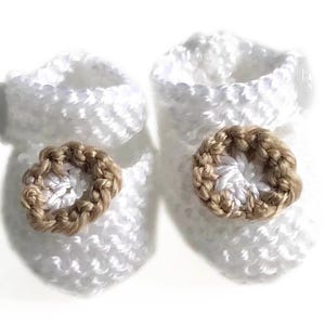 May include: A pair of white crochet baby booties. Each bootie features a brown, circular embellishment on the front. The booties are made of a soft, textured yarn, perfect for a newborn.