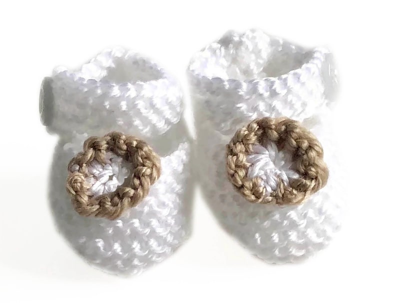 May include: A pair of white crochet baby booties. Each bootie features a brown, circular embellishment on the front. The booties are made of a soft, textured yarn, perfect for a newborn.