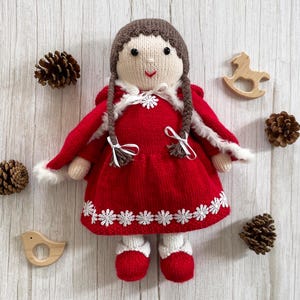 May include: A knitted doll wearing a red dress with white trim and a white fur-trimmed hooded cape. The doll has brown hair and a smiling face.