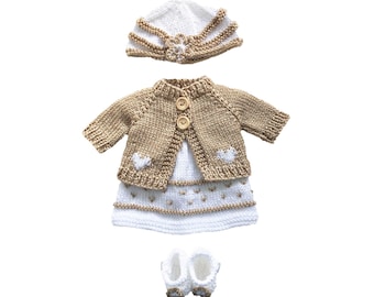 May include: A knitted doll outfit in white and tan. The set includes a hat, a cardigan with two wooden buttons, a dress with heart details, and a pair of booties. The outfit is designed for a doll and is made of soft yarn.