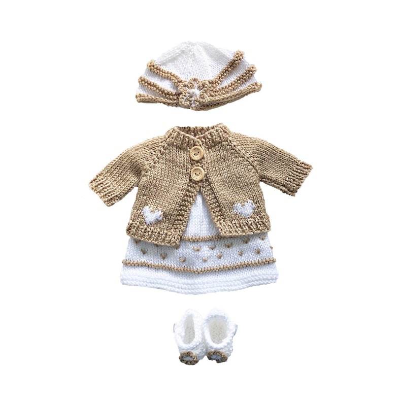 May include: A knitted doll outfit in white and tan. The set includes a hat, a cardigan with two wooden buttons, a dress with heart details, and a pair of booties. The outfit is designed for a doll and is made of soft yarn.