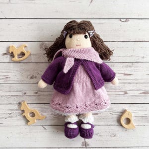 May include: A handmade knitted doll with brown yarn hair, wearing a purple jacket, scarf, and shoes. The doll has a pink dress and white socks. Wooden toys, including a rocking horse, dinosaur, and bird, are scattered around the doll.