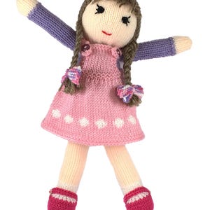 May include: A handmade knitted doll with brown braided hair, wearing a pink dress with white polka dots, purple sleeves, and pink shoes. The doll has a smiling face with black eyes and a red mouth. The arms are outstretched.
