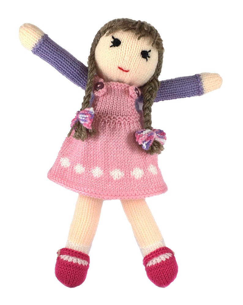 May include: A handmade knitted doll with brown braided hair, wearing a pink dress with white polka dots, purple sleeves, and pink shoes. The doll has a smiling face with black eyes and a red mouth. The arms are outstretched.