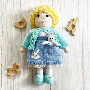 Pode incluir: Uma boneca feita à mão com cabelo loiro encaracolado, vestindo um vestido de malha azul com um desenho de coelho branco e um cardigan turquesa. A boneca tem sapatos turquesa e meias brancas. Brinquedos de madeira em forma de pássaro, urso e cavalo de balanço são colocados ao redor da boneca.