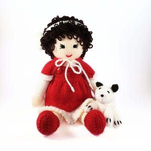 May include: A knitted doll with brown curly hair wearing a red dress and white boots. The doll is holding a white knitted dog with black ears.