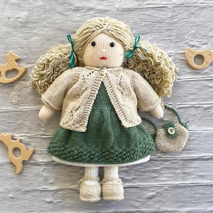 May include: A handmade doll with blonde yarn hair styled in pigtails, wearing a green dress and a cream-colored cardigan. The doll has a small purse and is accessorized with cream-colored shoes. Wooden toys are placed around the doll.