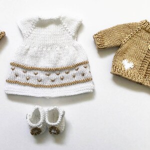 May include: A knitted doll clothing set in white and tan. The set includes a dress, cardigan, hat, and shoes. The dress and hat have tan accents. The cardigan has two wooden buttons and heart-shaped details. The shoes have tan accents.
