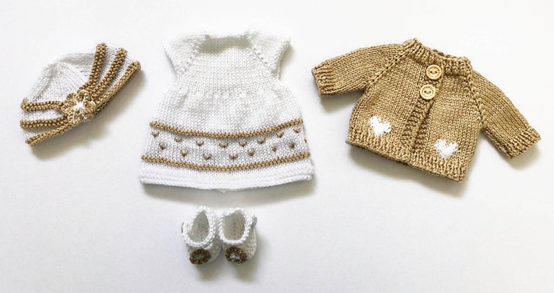 May include: A knitted doll clothing set in white and tan. The set includes a dress, cardigan, hat, and shoes. The dress and hat have tan accents. The cardigan has two wooden buttons and heart-shaped details. The shoes have tan accents.