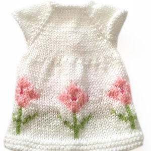 May include: A white knitted dress with short sleeves, featuring three pink tulip designs with green stems and leaves. The dress is handmade and has a textured appearance, perfect for a baby or toddler.