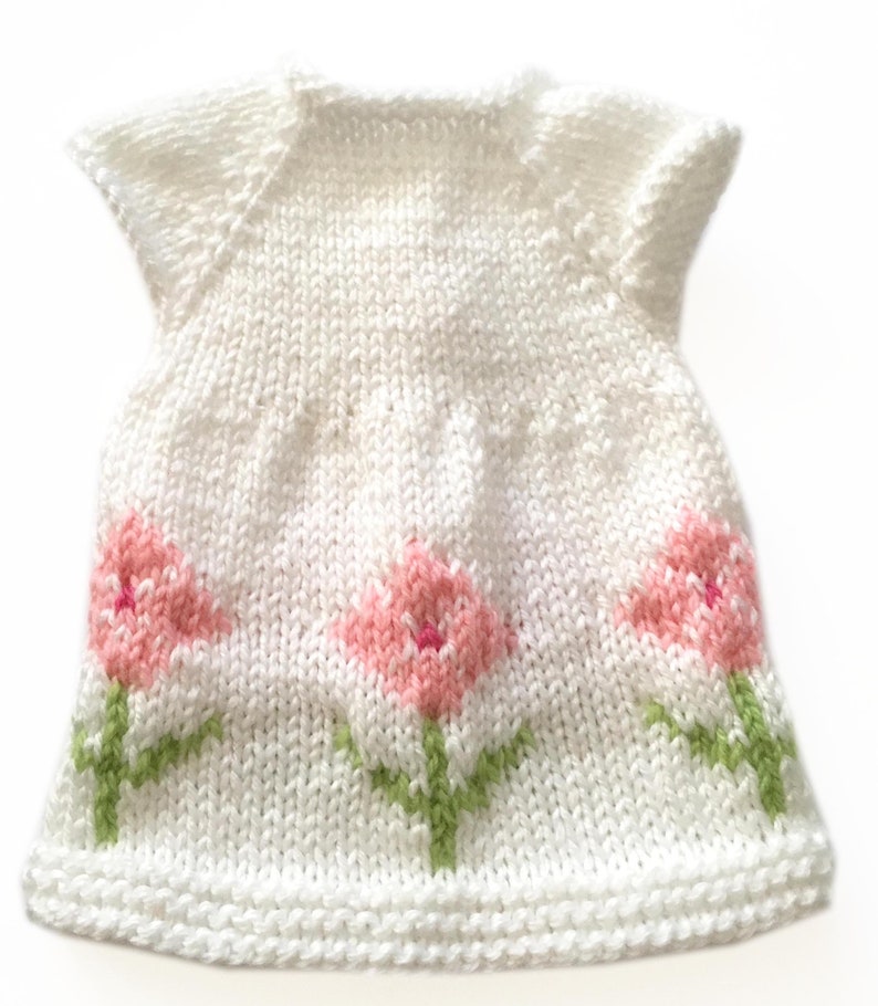 May include: A white knitted dress with short sleeves, featuring three pink tulip designs with green stems and leaves. The dress is handmade and has a textured appearance, perfect for a baby or toddler.