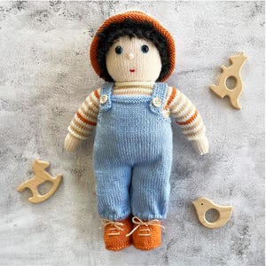 May include: A knitted doll with brown hair, wearing a blue and white striped shirt, blue overalls, and orange shoes. The doll is surrounded by wooden toys.