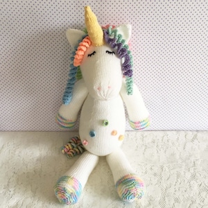 May include: A handmade white knitted unicorn stuffed animal with a yellow horn, rainbow mane, and tail. The unicorn has closed eyes, and colorful accents on its body and feet. The toy is sitting on a white lace surface.