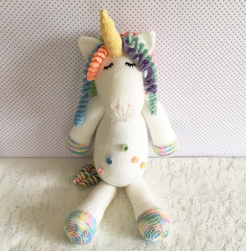 May include: A handmade white knitted unicorn stuffed animal with a yellow horn, rainbow mane, and tail. The unicorn has closed eyes, and colorful accents on its body and feet. The toy is sitting on a white lace surface.