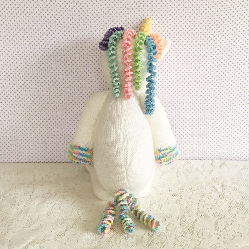 May include: A handmade white knitted unicorn plush toy with a yellow horn, rainbow mane, and tail. The unicorn has pastel rainbow accents on the hooves and sleeves. The toy is sitting on a white lace surface.