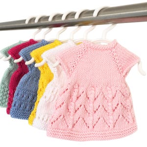 Pode incluir: Uma fileira de seis vestidos de bebê de malha coloridos pendurados em cabides brancos. Os vestidos são em tons de verde, rosa, azul, amarelo, branco e rosa. Cada vestido tem um design único, com padrões texturizados e mangas curtas.