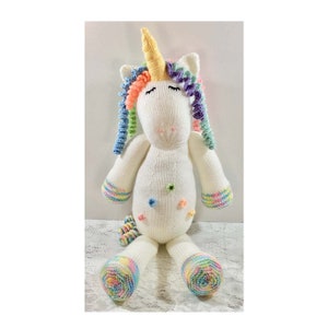 May include: A handmade, white knitted unicorn plush toy with a yellow horn, rainbow mane, and tail. The unicorn has closed eyes, colorful accents, and small, decorative flower-shaped buttons. The hooves and cuffs of the arms are also decorated with rainbow stripes.