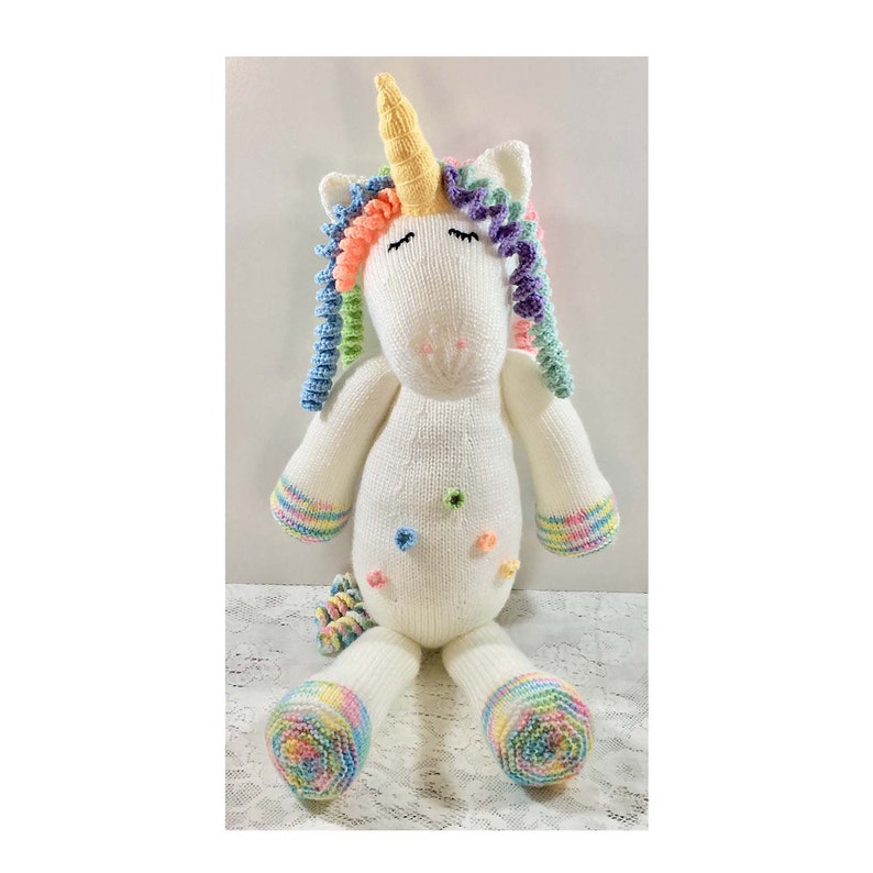 May include: A handmade, white knitted unicorn plush toy with a yellow horn, rainbow mane, and tail. The unicorn has closed eyes, colorful accents, and small, decorative flower-shaped buttons. The hooves and cuffs of the arms are also decorated with rainbow stripes.