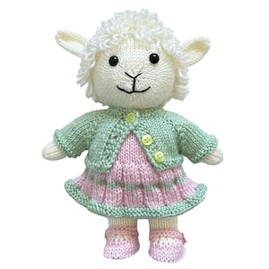 Op de afbeelding: Een handgebreid knuffellammetje van crèmekleurige wol met zwarte kraaloogjes en een geborduurde neus. Het lammetje draagt een lichtgroen gebreid vestje met drie kleine gele knoopjes over een roze en groen gestreept jurkje en bijpassende roze schoentjes.