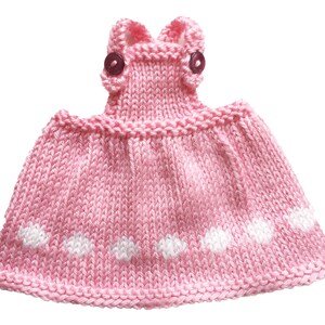 May include: A handmade pink knitted dress with white polka dots. The dress features a detailed bodice with two small, dark buttons and a flared skirt. The knitted fabric has a textured appearance, and the dress is designed for a small child or doll.