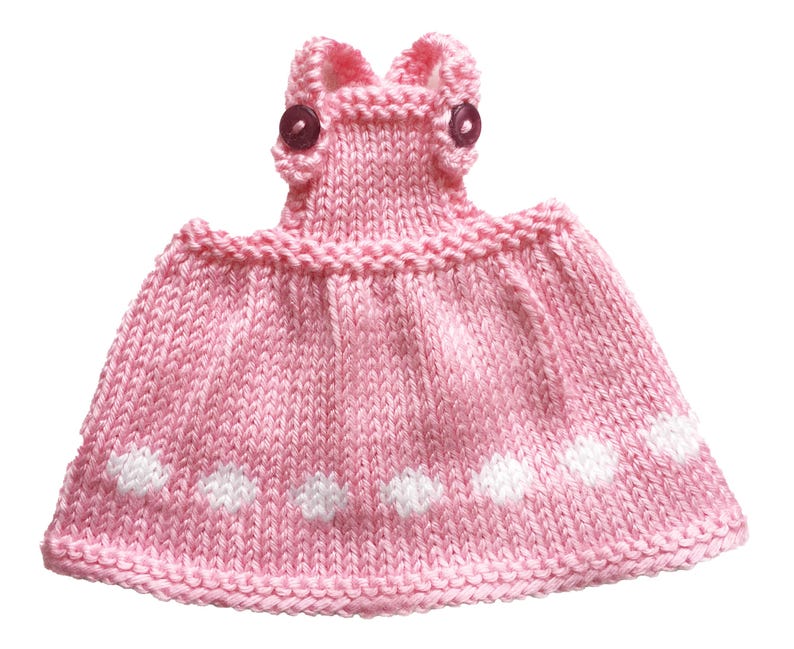 May include: A handmade pink knitted dress with white polka dots. The dress features a detailed bodice with two small, dark buttons and a flared skirt. The knitted fabric has a textured appearance, and the dress is designed for a small child or doll.
