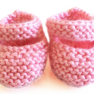 May include: A pair of handmade baby booties in a soft pink hue. These knitted booties feature a folded cuff and a small button detail on the side. The cozy, textured knit suggests warmth and comfort for a newborn.