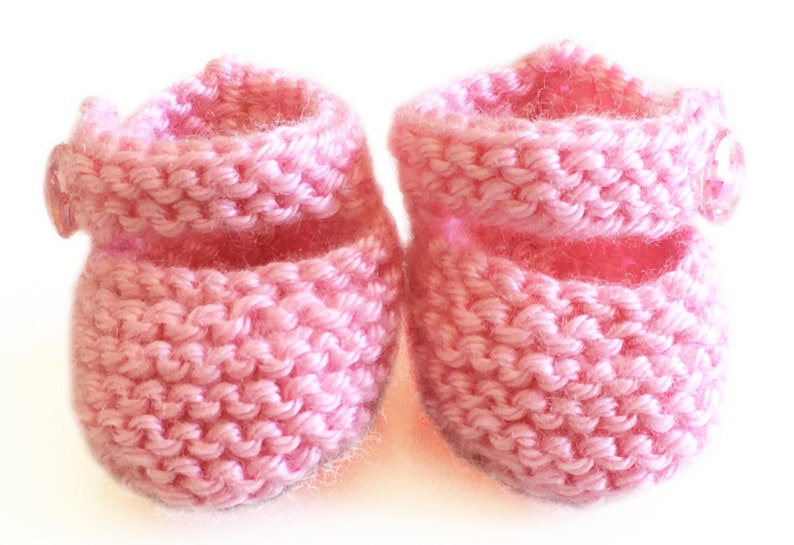 May include: A pair of handmade baby booties in a soft pink hue. These knitted booties feature a folded cuff and a small button detail on the side. The cozy, textured knit suggests warmth and comfort for a newborn.