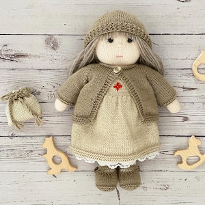 Op de afbeelding: Een gebreide pop met lang blond haar, die een bruine cardigan, een beige jurk en een bruine gebreide muts draagt. De pop houdt een klein bruin tasje in haar hand.